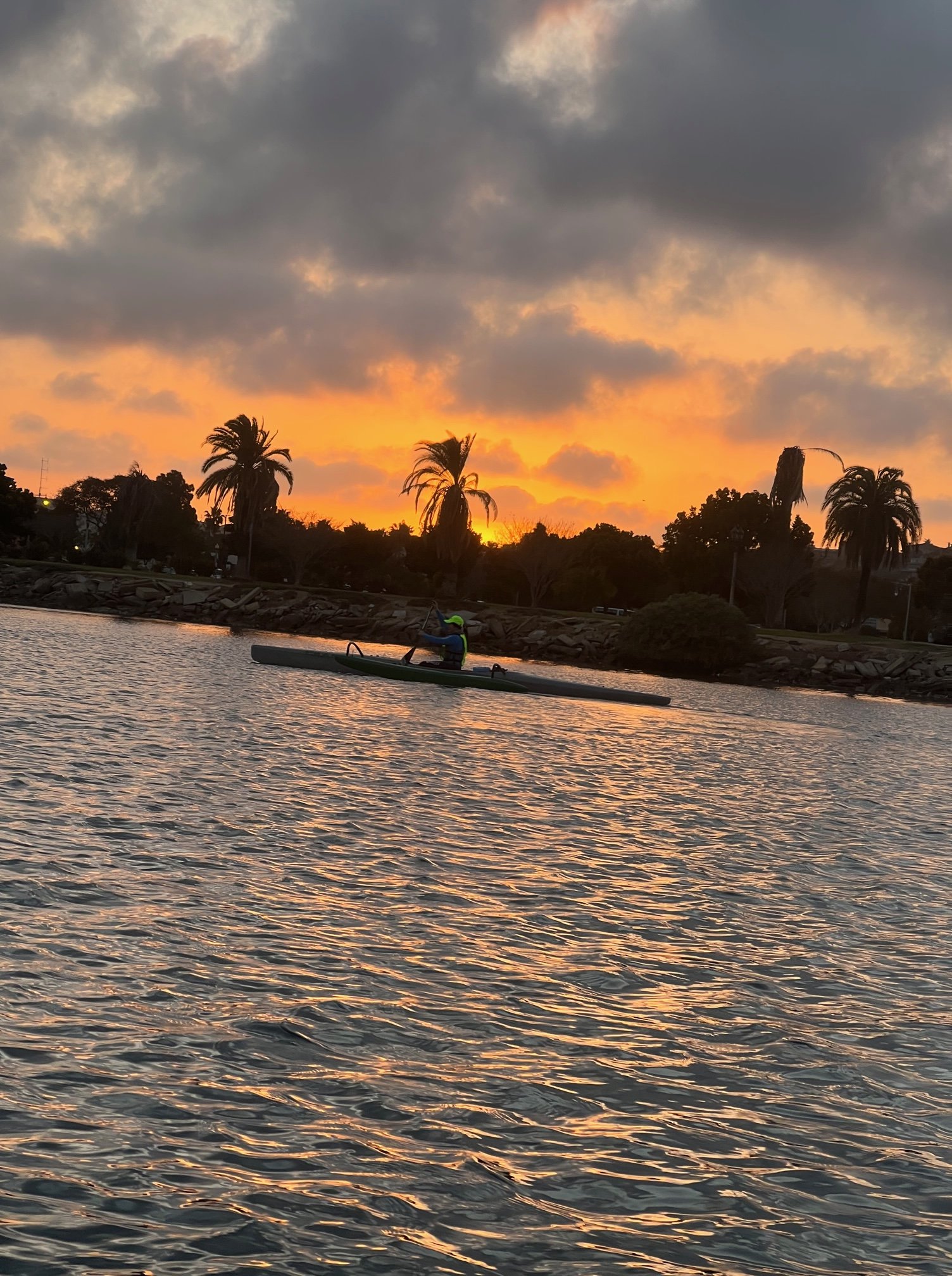 A paddler on an OC1, and in the background a cloudy sunset behind a dark shore lined with palm trees.